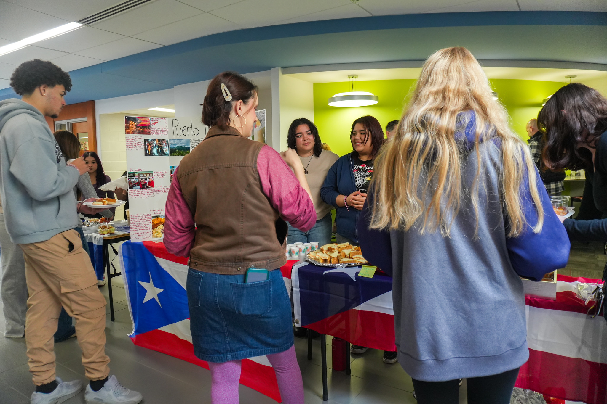 Students gather around a Puerto Rico-themed display table decorated with the Puerto Rican flag and traditional foods during a humanities week cultural fair event on a college campus