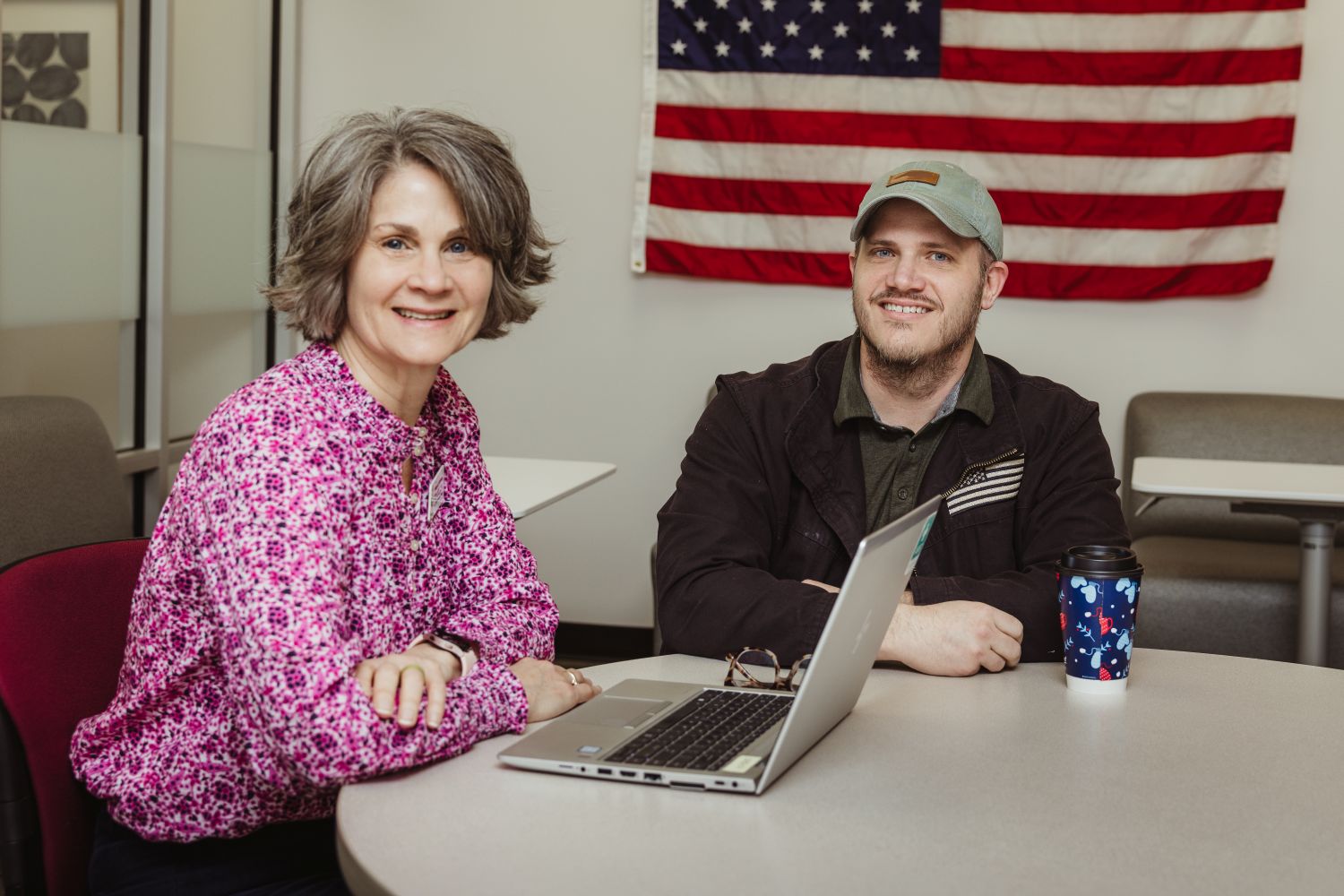 Sharon Painter sits at a table with a smiling veteran student with an American flag in the background.