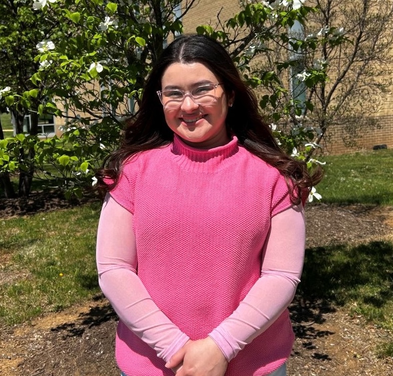 A young woman with long dark hair and clear-framed glasses smiles while standing outdoors on a sunny day, wearing a pink long-sleeve shirt layered under a pink knit sweater vest, with a blooming dogwood tree and a brick campus building visible in the background.