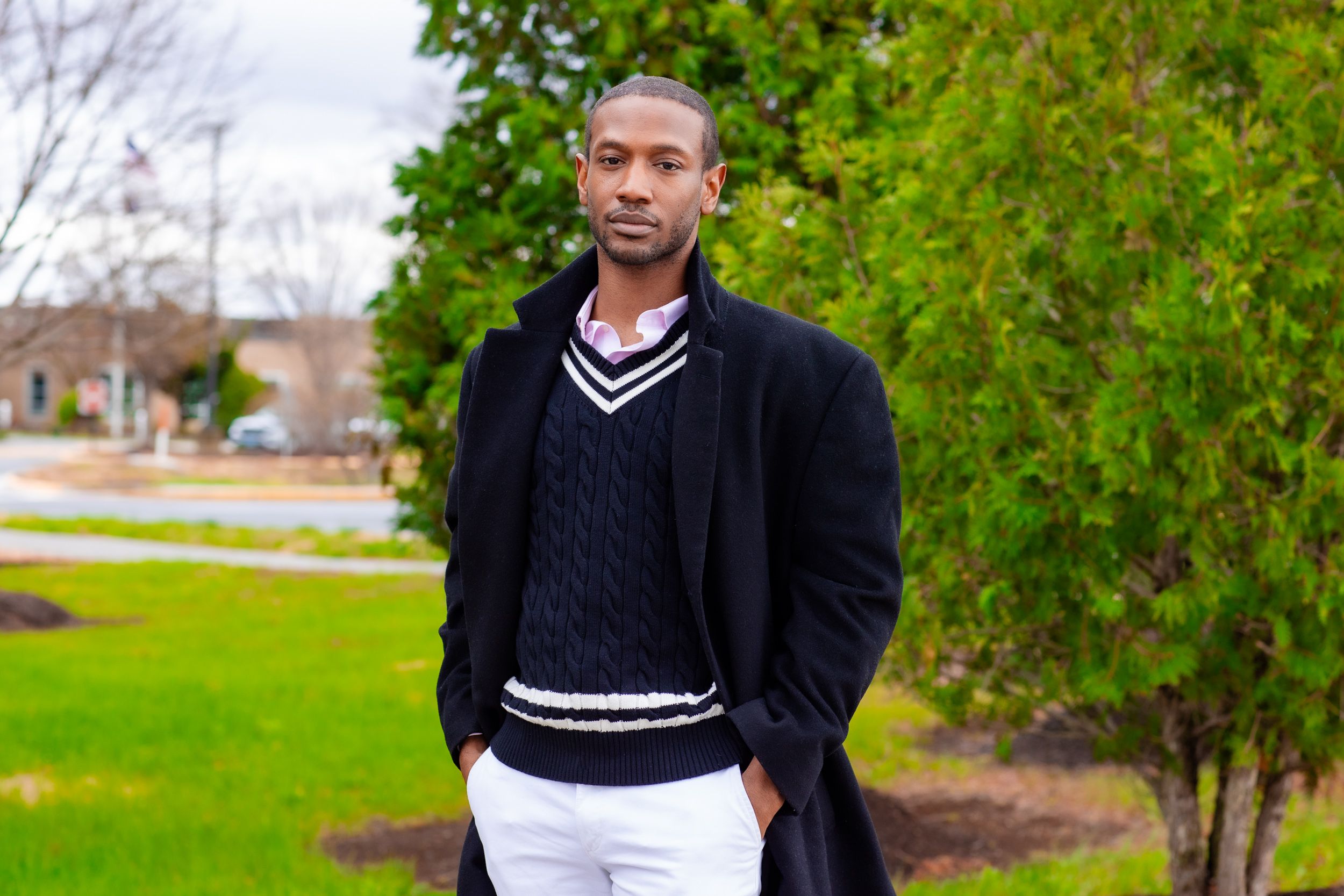 Jamal Plummer stands outdoors on a green lawn, wearing a black cable-knit v-neck sweater with white stripe trim layered over a pink collared shirt and under a long black coat, with white pants. He faces the camera with a composed expression, hands in his pockets. Green shrubs and trees are visible in the background, along with a blurred street scene and overcast sky.