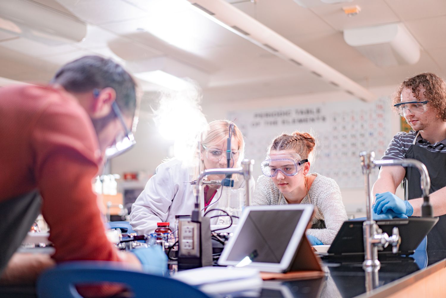 A Laurel Ridge Community College instructor in a white lab coat and safety goggles works alongside three students at a chemistry lab bench. The students wear safety goggles and blue nitrile gloves as they observe an experiment producing visible vapor or steam. Lab equipment, small reagent bottles, and tablets are arranged on the bench. A periodic table of the elements is visible on the wall in the background.
