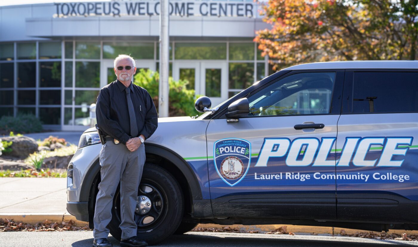 A man with white hair and a beard, wearing sunglasses, a dark button-up shirt, tie, and gray pants, stands beside a Laurel Ridge Community College Police patrol vehicle in front of the Toxopeus Welcome Center on a sunny autumn day.