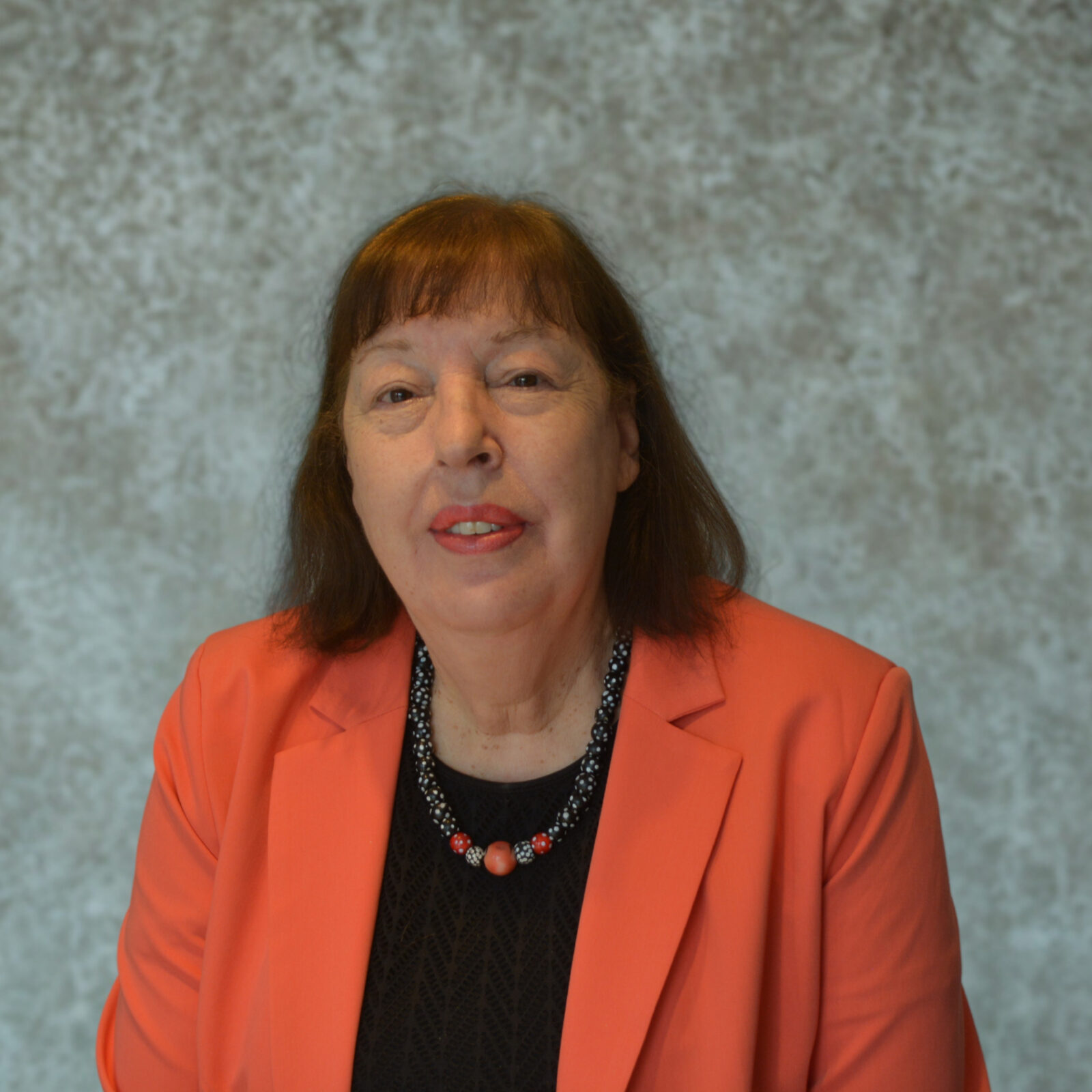 Head-and-shoulders portrait of Virginia Gamba with brown hair and bangs, wearing a coral blazer, black top, and beaded necklace, seated against a gray textured backdrop.