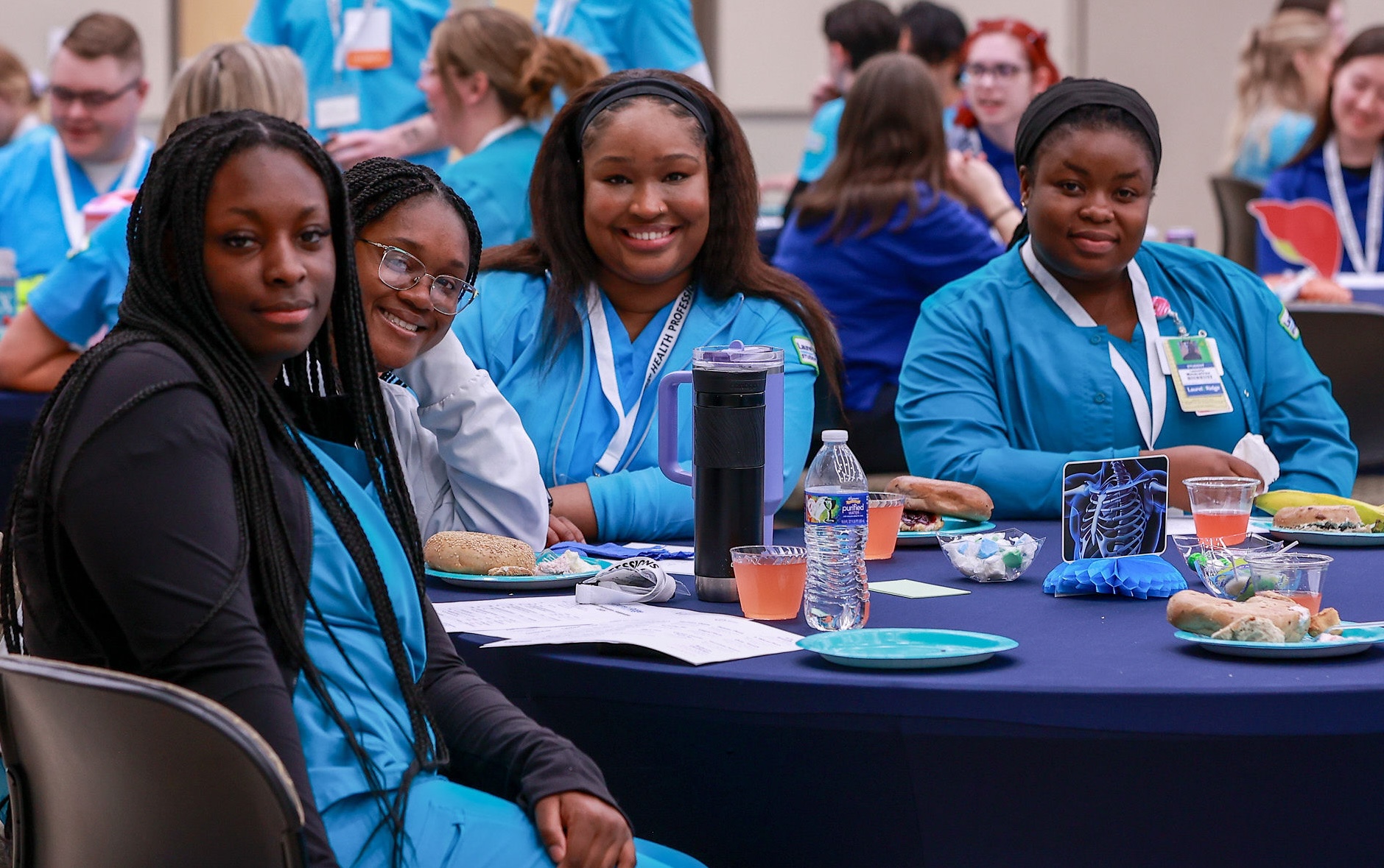 Four students wearing teal scrubs and lanyards smile at the camera while seated at a round table with a navy blue tablecloth during a health professions symposium luncheon.