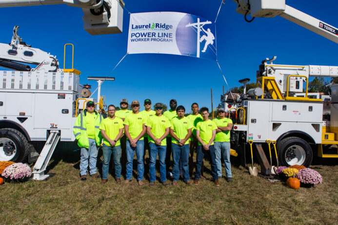 A group of power line worker graduates stand in front of two power line worker vehicles.
