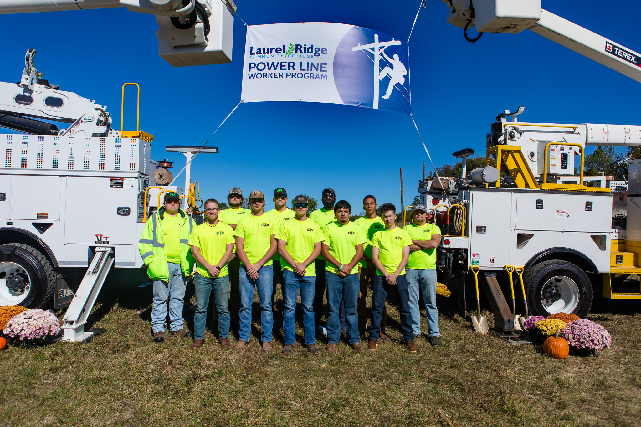 A group of power line worker graduates stand in front of two power line worker vehicles.
