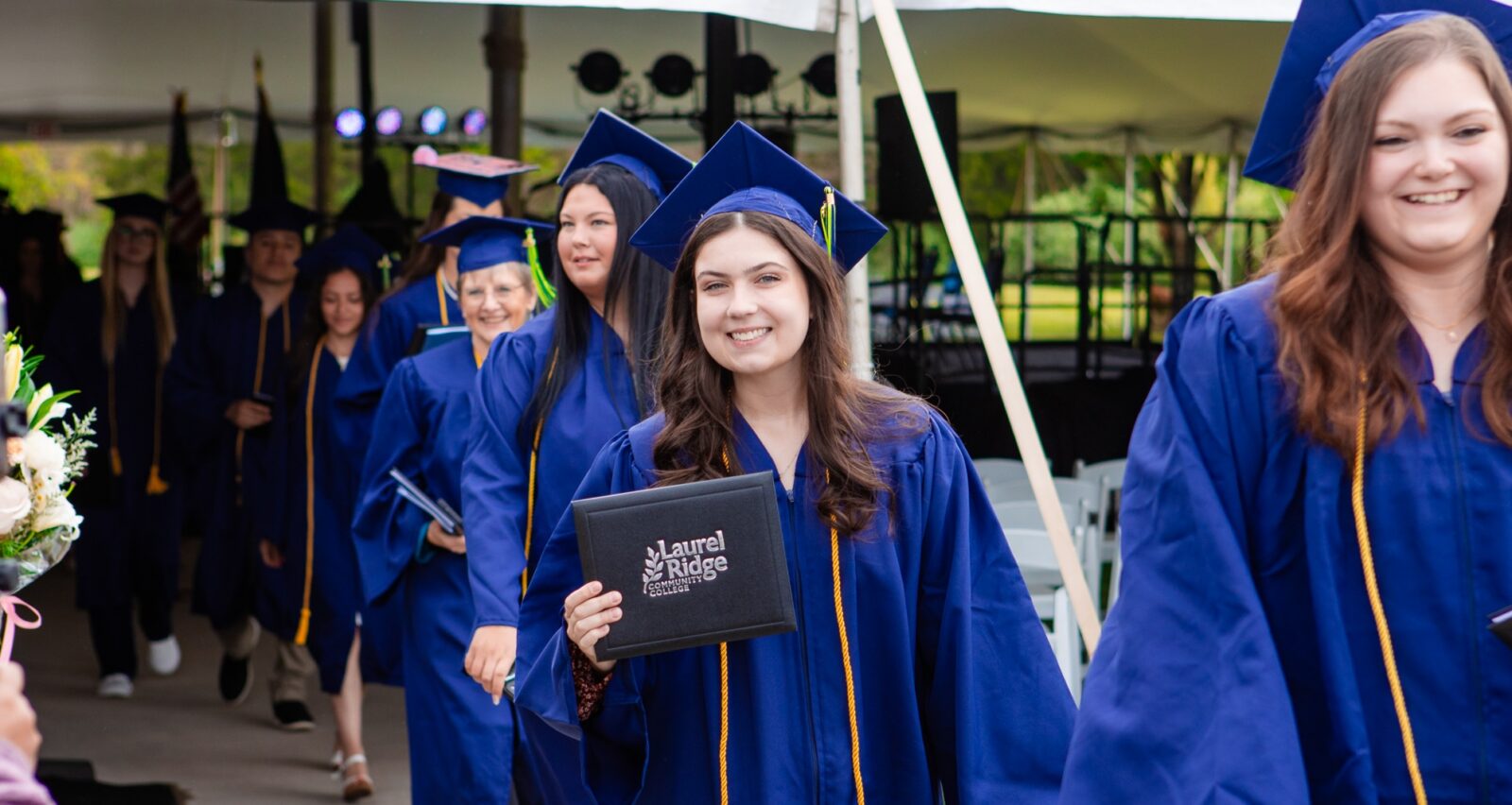 A smiling Laurel Ridge Community College graduate in a blue cap and gown holds her diploma cover while processing with fellow graduates during the outdoor commencement ceremony.