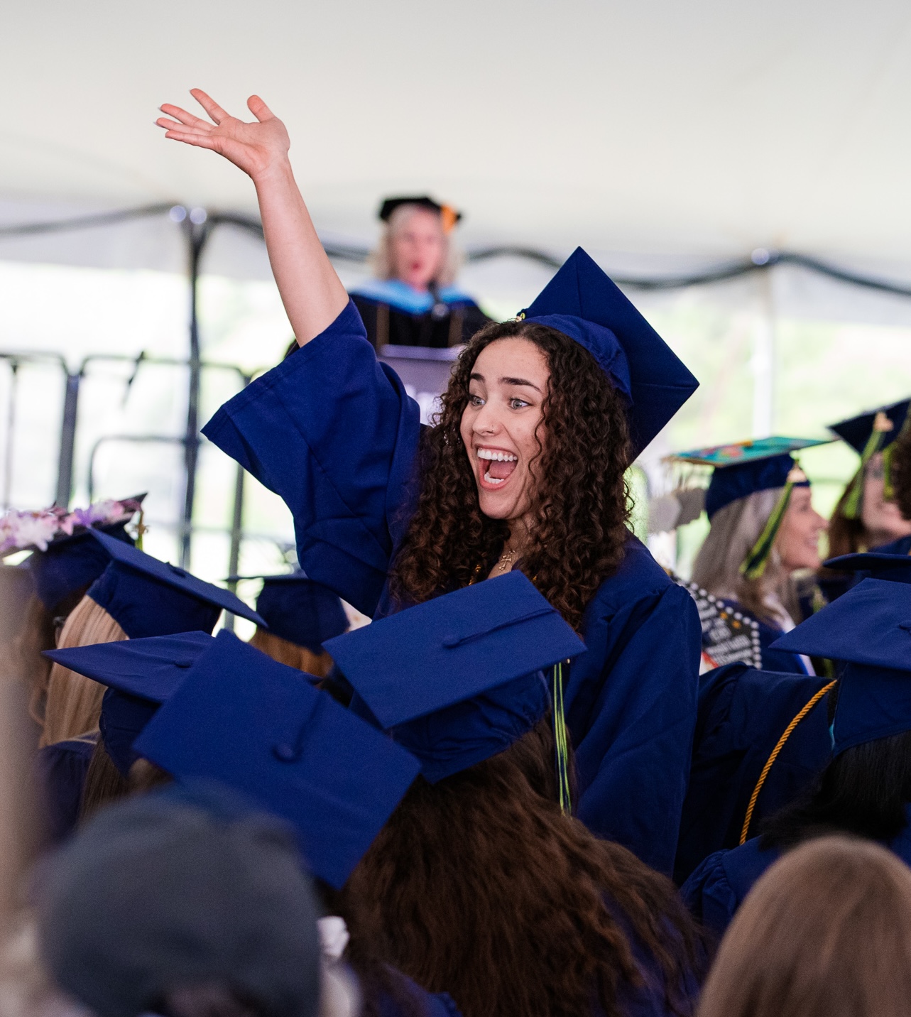 A Laurel Ridge Community College graduate in a blue cap and gown raises her hand and cheers with excitement while seated among fellow graduates during the commencement ceremony.