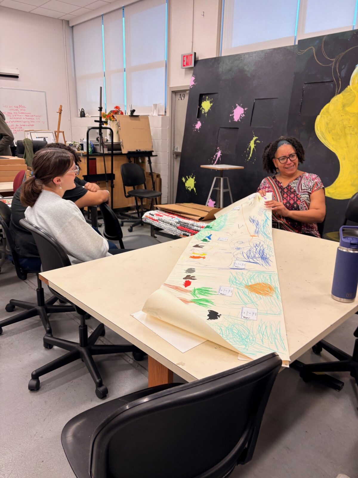 Professor Monica James standing with FEA students inside the art classroom during exhibit preparation.