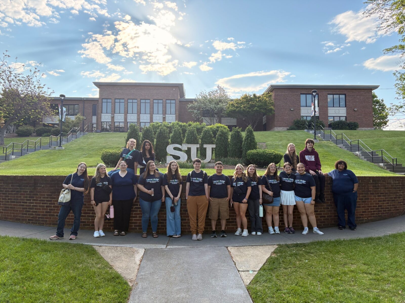 A group of approximately 18 students pose in front of a large "STU" letter sign on a landscaped brick retaining wall, with a brick academic building visible in the background. Most students wear matching navy blue Laurel Ridge Future Educators Academy t-shirts. The group is arranged in two informal rows, with some students standing along the wall and others seated on top of it. The photo is taken on a sunny day with a bright sky and scattered clouds.