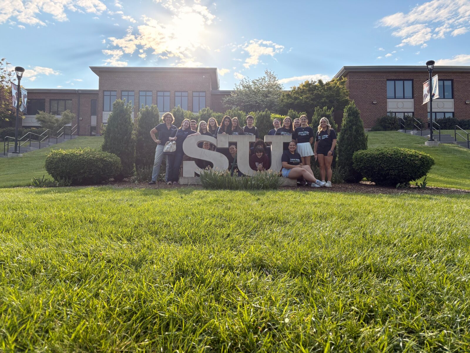 FEA students and their director standing together in front of the Shenandoah University campus sign.
