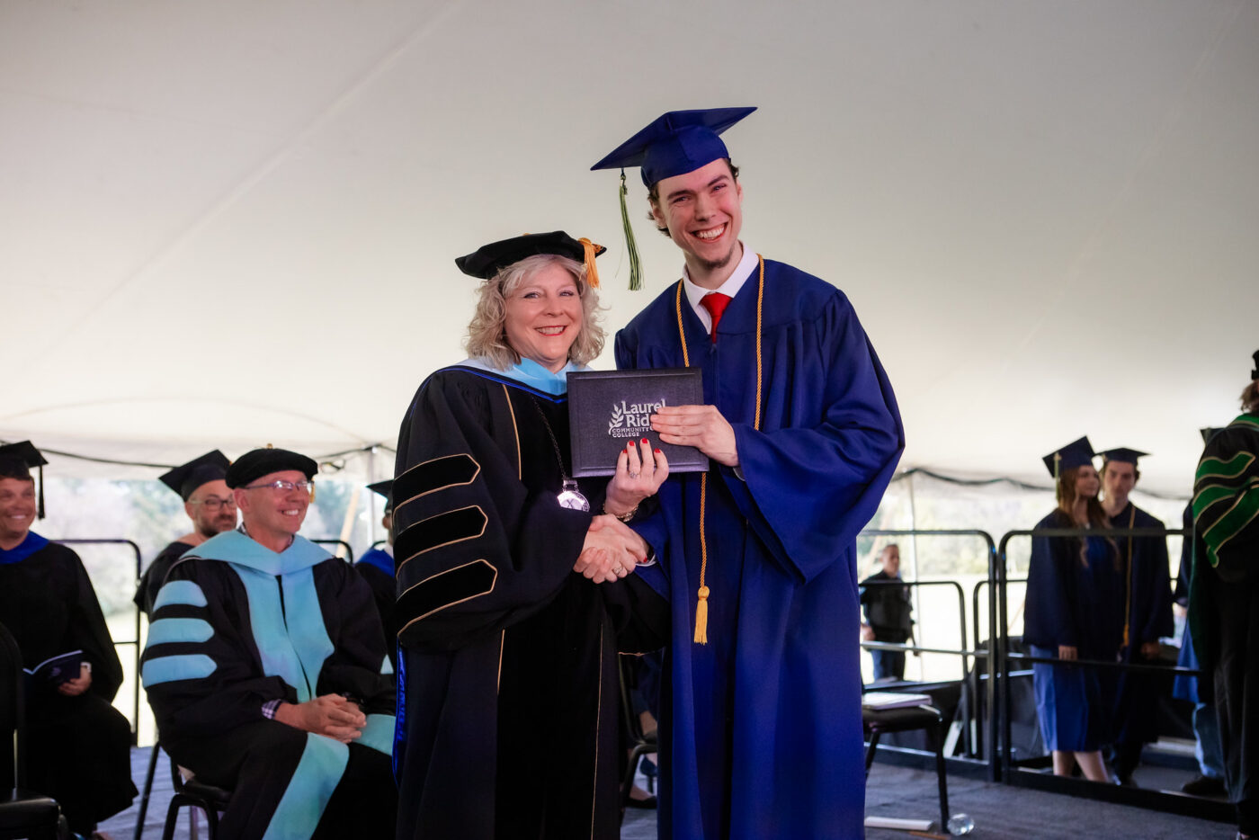 Judah Dotson smiles while receiving his diploma folder from a Laurel Ridge Community College administrator during commencement ceremonies held under a large tent. Dotson wears a blue cap and gown with gold and green honor cords and a red tie, while the administrator is dressed in black doctoral regalia with blue and gold accents. Faculty and fellow graduates are visible in the background.