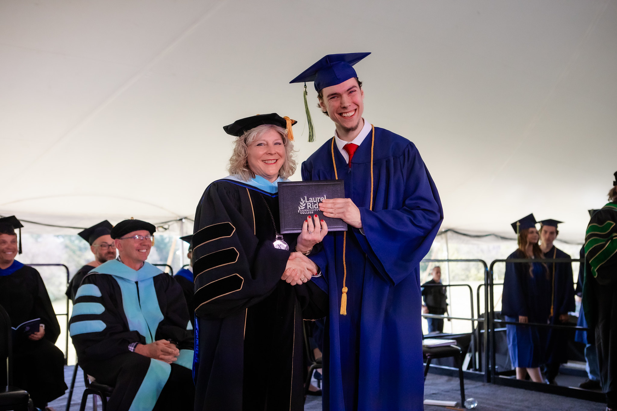 Judah Dotson smiles while receiving his diploma folder from a Laurel Ridge Community College administrator during commencement ceremonies held under a large tent. Dotson wears a blue cap and gown with gold and green honor cords and a red tie, while the administrator is dressed in black doctoral regalia with blue and gold accents. Faculty and fellow graduates are visible in the background.