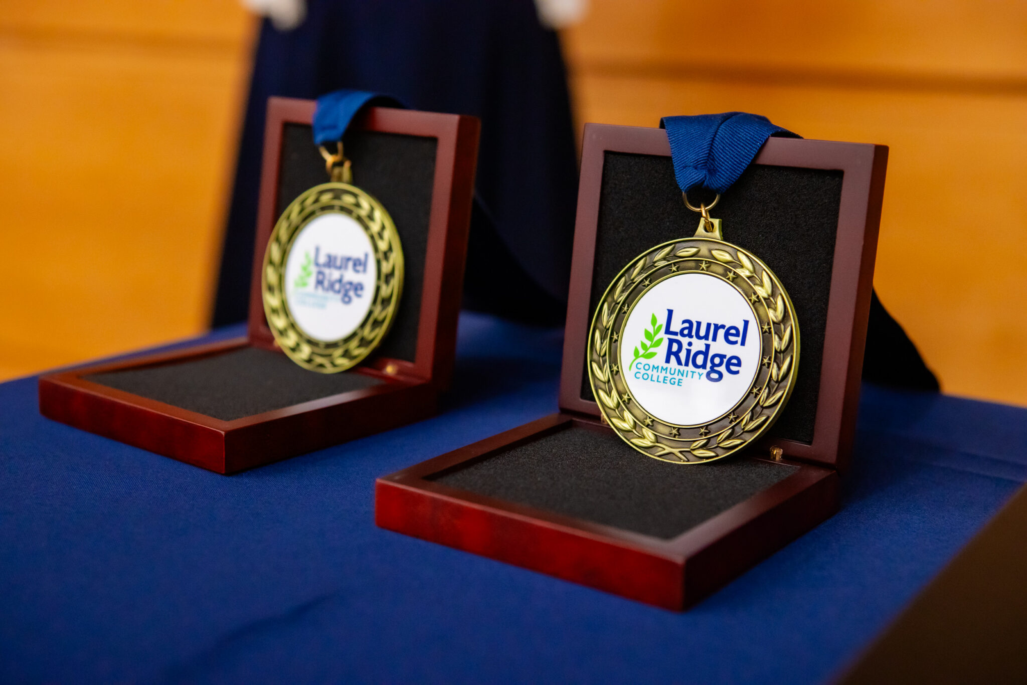 Two Laurel Ridge Community College Medallions of Recognition rest in open wooden presentation boxes on a blue-draped table. The gold medals feature the college's logo and laurel wreath design, each with a blue ribbon.