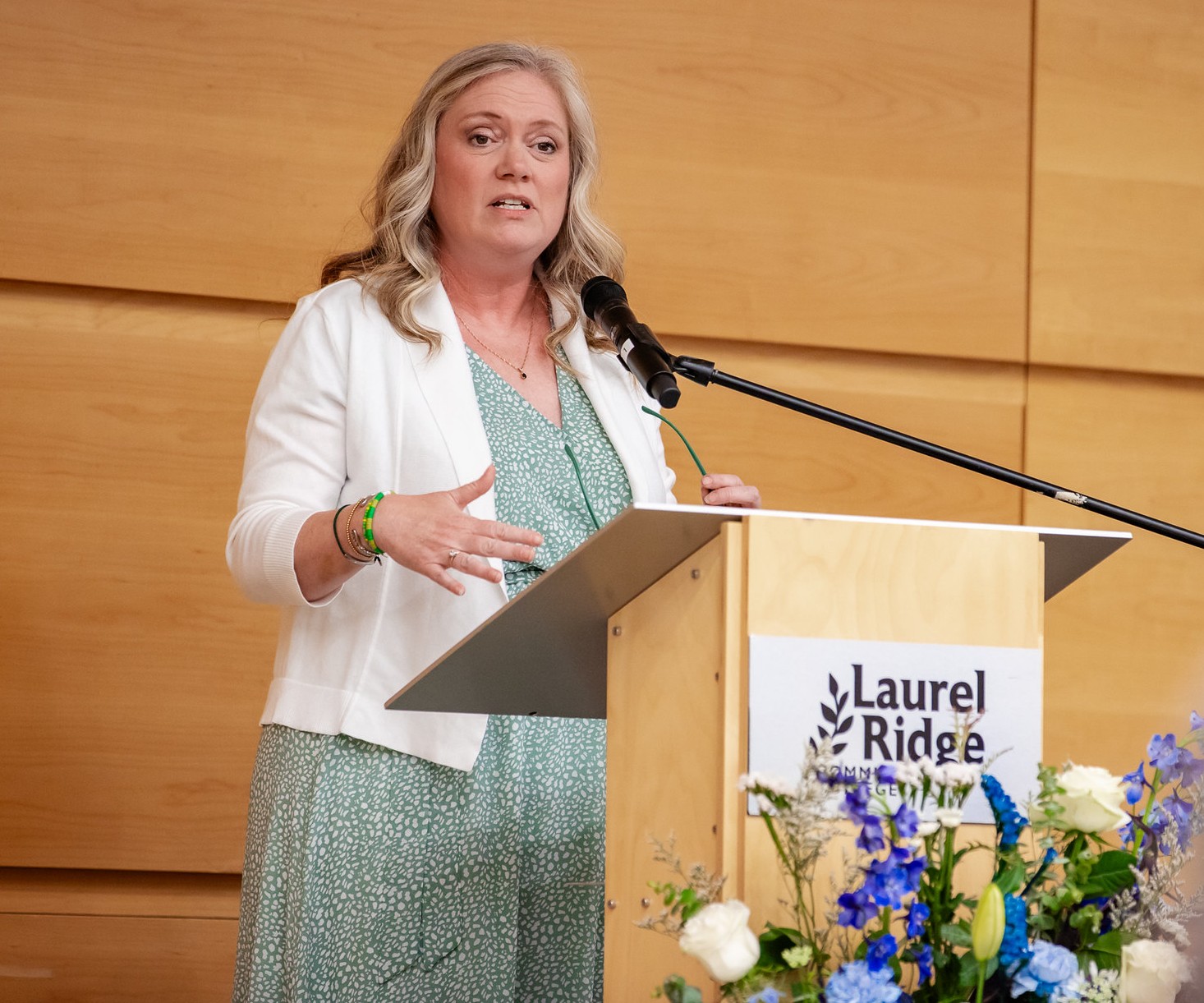 Crystal Lynn speaks at a Laurel Ridge Community College podium during the 2026 Medallion of Recognition ceremony. She is wearing a white cardigan over a green floral dress and gestures with her hand while addressing the audience.
