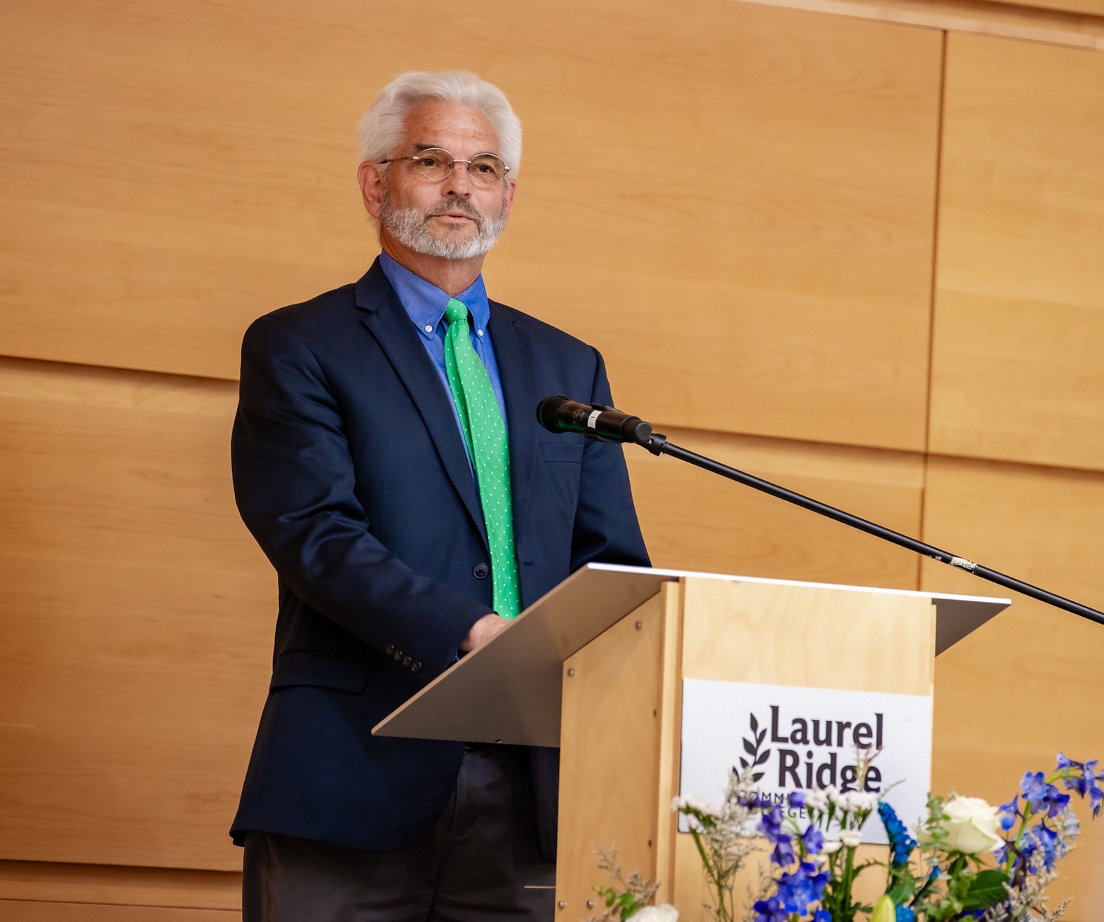 Larry Baker speaks at a Laurel Ridge Community College podium during the 2026 Medallion of Recognition ceremony. He is wearing a navy blazer, blue shirt, and green tie, with white hair and glasses.
