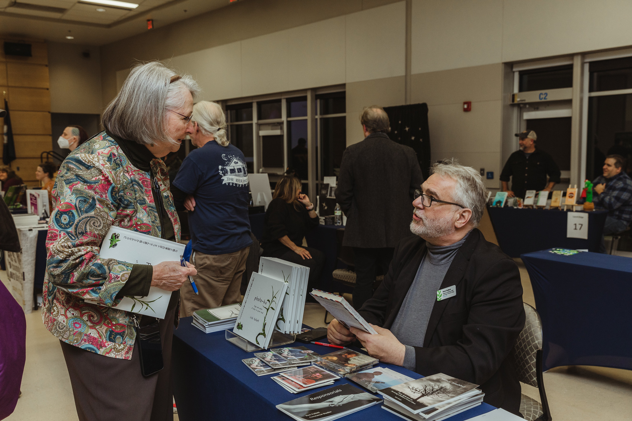 A woman in a colorful floral jacket speaks with a male author seated at a book fair vendor table displaying copies of his book "photo-ku" and other publications, during the Laurel Ridge Community College Book Fair 2022.