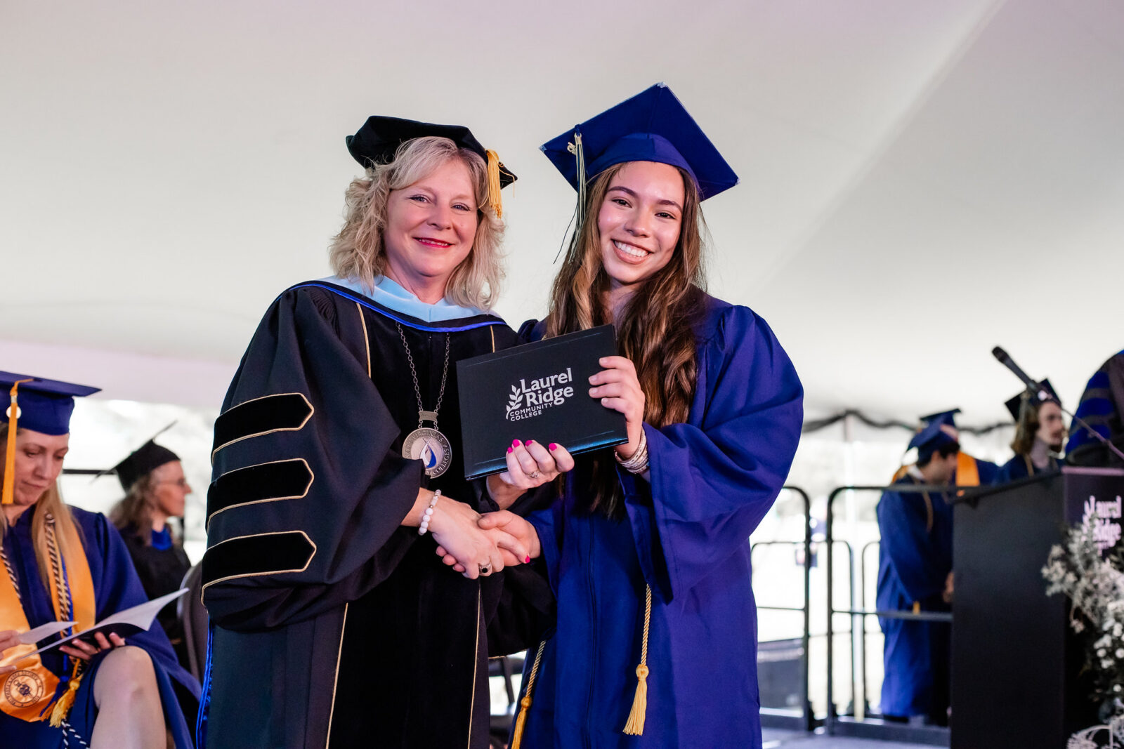 A Laurel Ridge Community College administrator in black doctoral regalia with gold trim and a medallion shakes hands with a smiling graduate in a blue cap and gown as they jointly hold a black diploma cover bearing the Laurel Ridge Community College logo. Other graduates and faculty are visible in the background beneath a large white tent.
