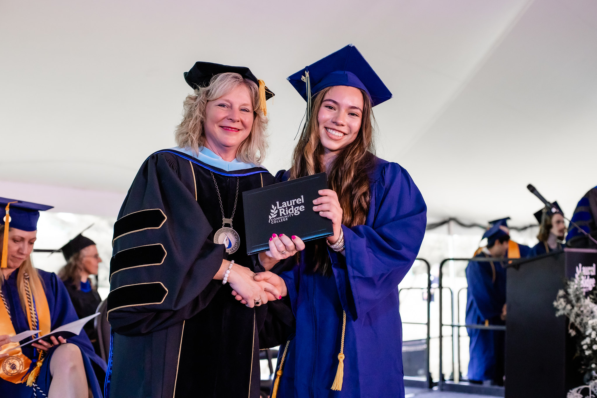 A Laurel Ridge Community College administrator in black doctoral regalia with gold trim and a medallion shakes hands with a smiling graduate in a blue cap and gown as they jointly hold a black diploma cover bearing the Laurel Ridge Community College logo. Other graduates and faculty are visible in the background beneath a large white tent.