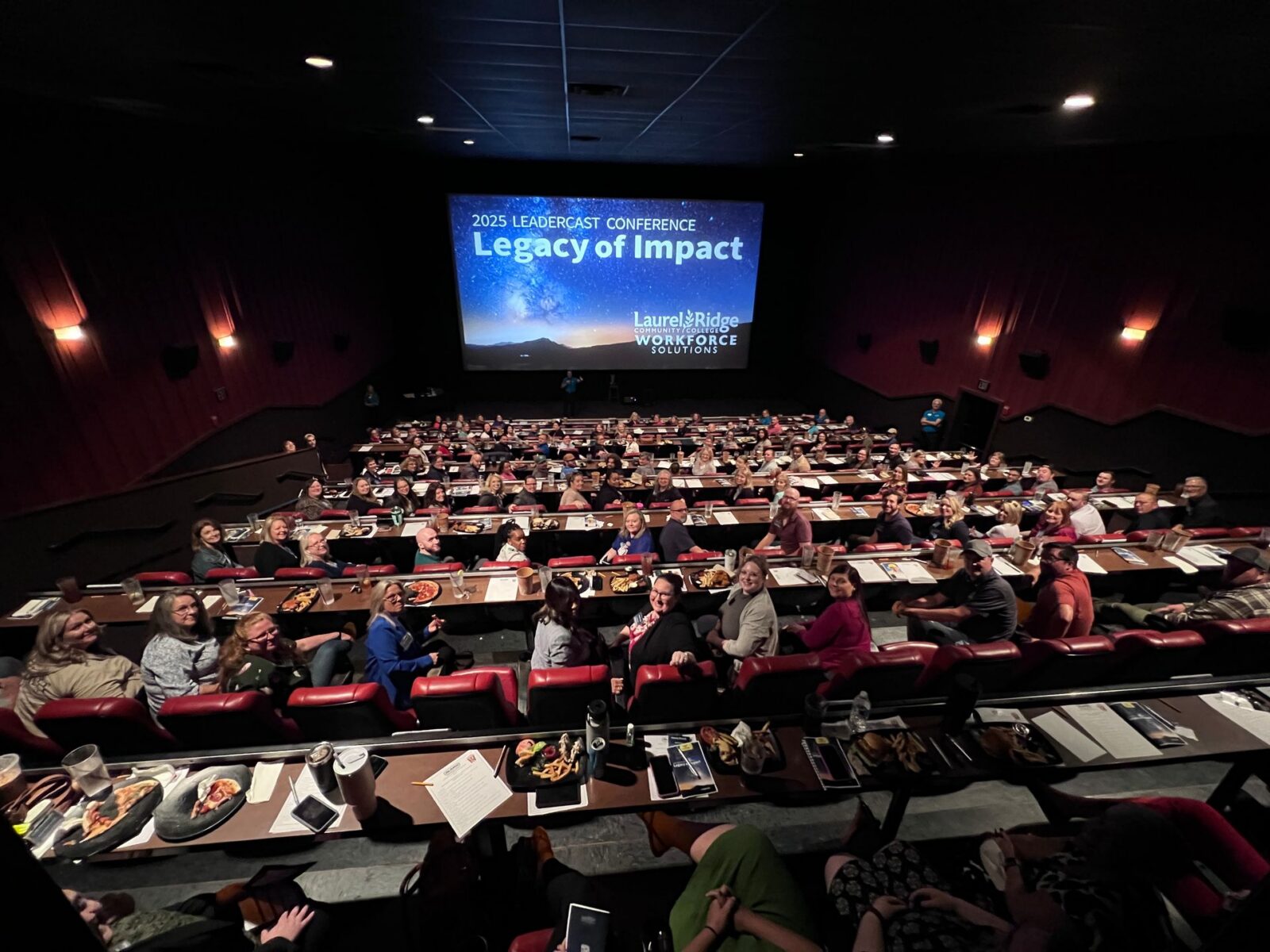 A large audience of attendees seated in a dine-in movie theater during the 2025 Leadercast Conference hosted by Laurel Ridge Community College Workforce Solutions. Guests seated at tiered rows of tables with food and drinks look toward a large screen displaying the conference theme "Legacy of Impact" against a starry night sky backdrop.