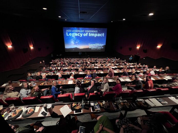 A large audience of attendees seated in a dine-in movie theater during the 2025 Leadercast Conference hosted by Laurel Ridge Community College Workforce Solutions. Guests seated at tiered rows of tables with food and drinks look toward a large screen displaying the conference theme "Legacy of Impact" against a starry night sky backdrop.