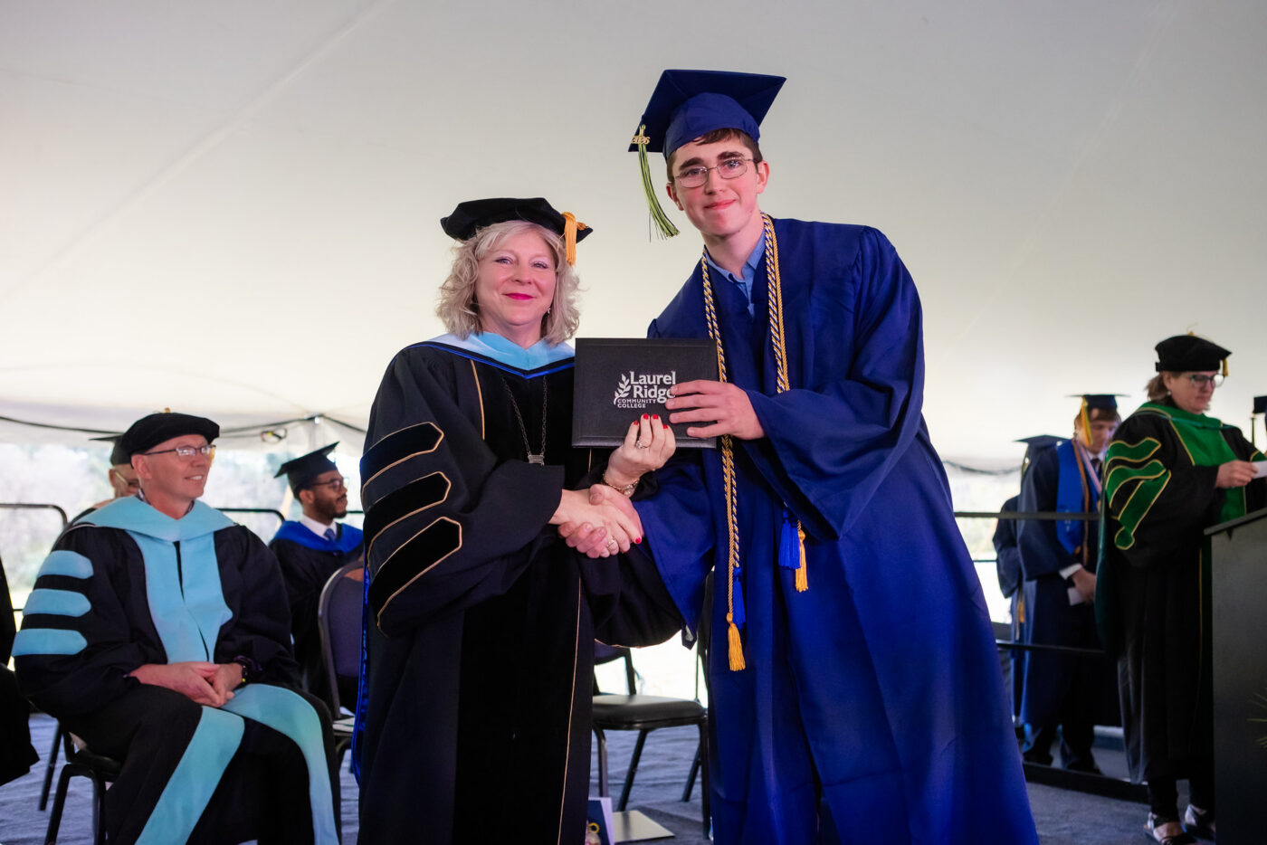 A Laurel Ridge Community College administrator in black doctoral regalia with light blue hood and gold trim shakes hands with a graduate in a blue cap and gown adorned with gold and blue honor cords, as the graduate receives a diploma portfolio bearing the Laurel Ridge Community College logo during an outdoor commencement ceremony held under a white tent.