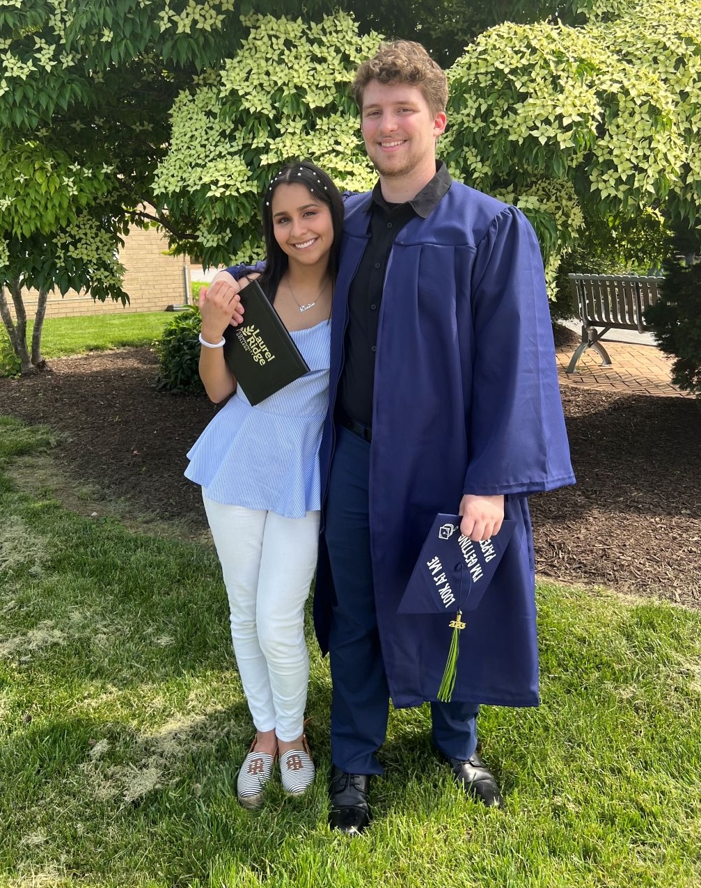 A young man in a navy blue graduation gown holds his cap with a green tassel while standing on a grassy lawn with his arm around a young woman wearing a light blue top and white pants. She holds a black Laurel Ridge Community College gift bag and smiles at the camera. Lush green trees and a brick building are visible in the background.