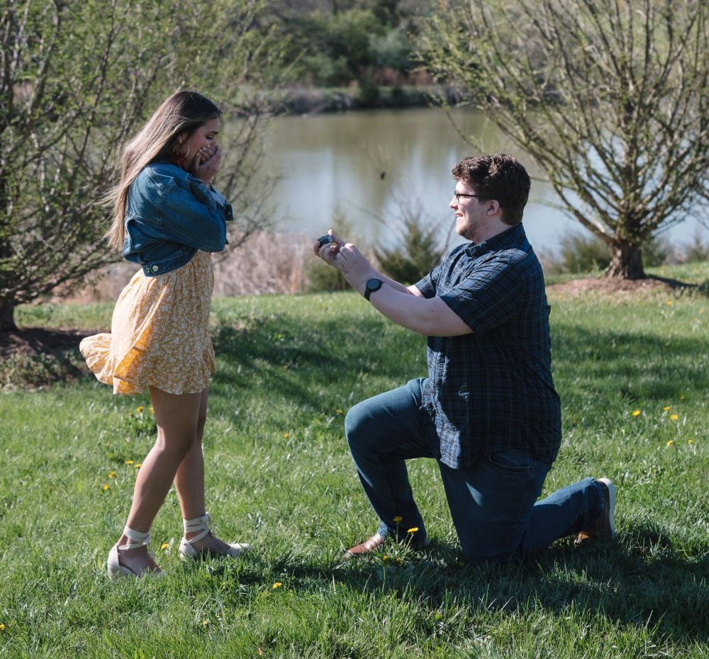 **Image 1:** A young man in a navy blue graduation gown holds his cap with a green tassel while standing on a grassy lawn with his arm around a young woman wearing a light blue top and white pants. She holds a black Laurel Ridge Community College gift bag and smiles at the camera. Lush green trees and a brick building are visible in the background. --- **Image 2:** A young man kneels on one knee on a grass lawn beside a pond, smiling as he holds open a ring box toward a young woman in a yellow floral dress and denim jacket, who covers her mouth with her hand in surprised delight. Budding trees and a calm body of water are visible in the background on a clear spring day.