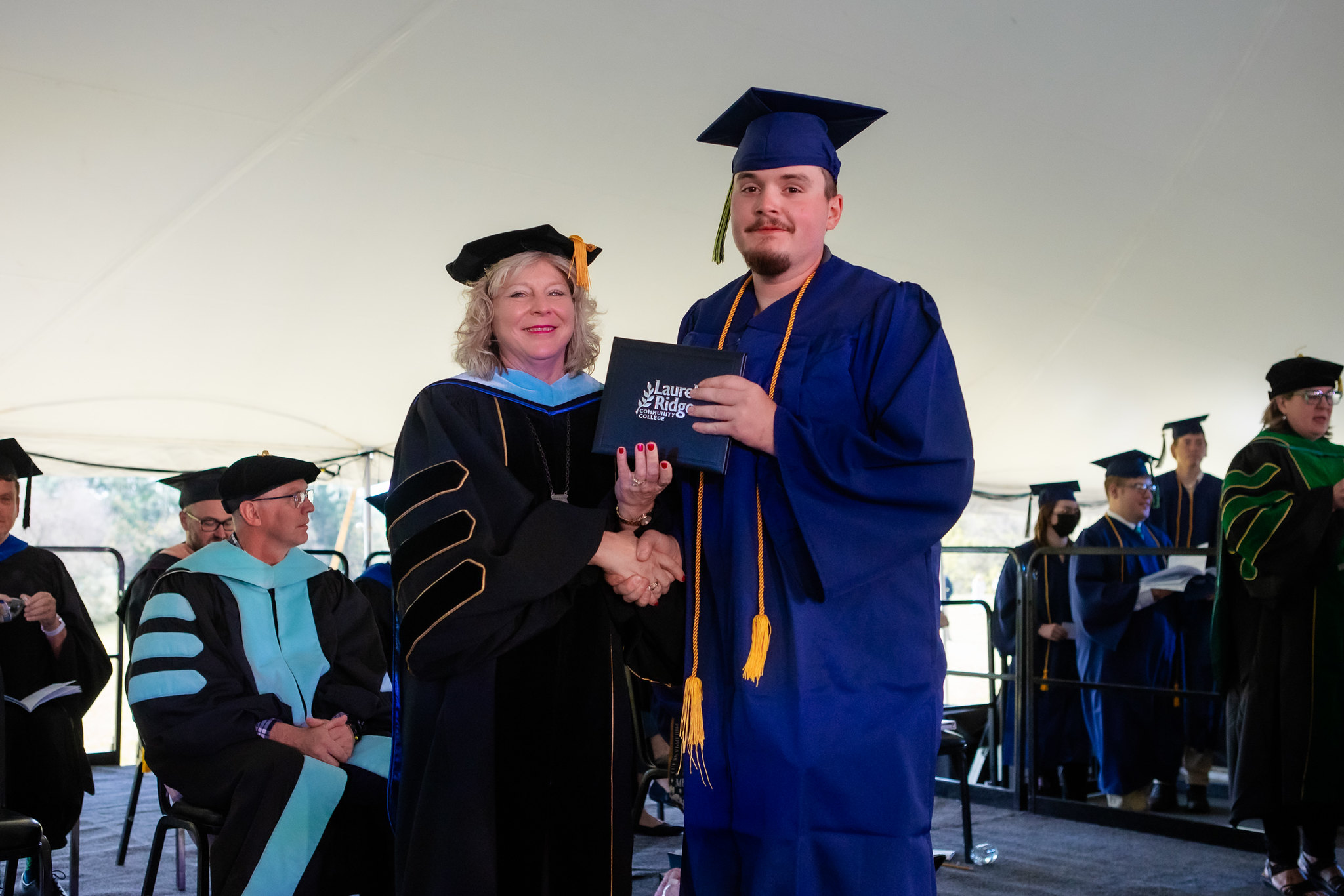 John Frazier, dressed in a blue cap and gown with a gold honor cord, shakes hands with a college administrator in black doctoral regalia with gold accents as he receives his Laurel Ridge Community College diploma cover. Both are smiling during the outdoor commencement ceremony held under a large tent, with faculty and fellow graduates visible in the background.