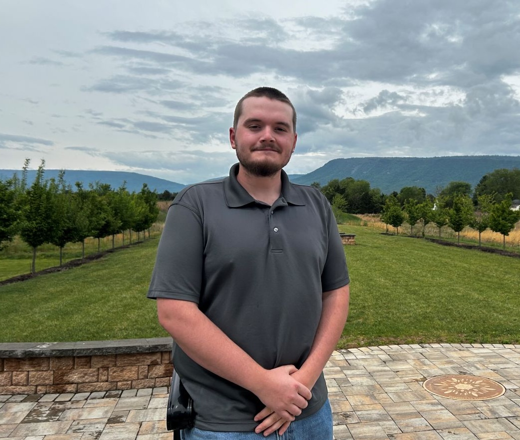 John Frazier stands outdoors on a paved stone patio, smiling at the camera with his hands clasped in front of him. He is wearing a gray polo shirt and jeans. Behind him, a landscaped lawn flanked by rows of young trees leads to a mountain ridge under a cloudy sky.