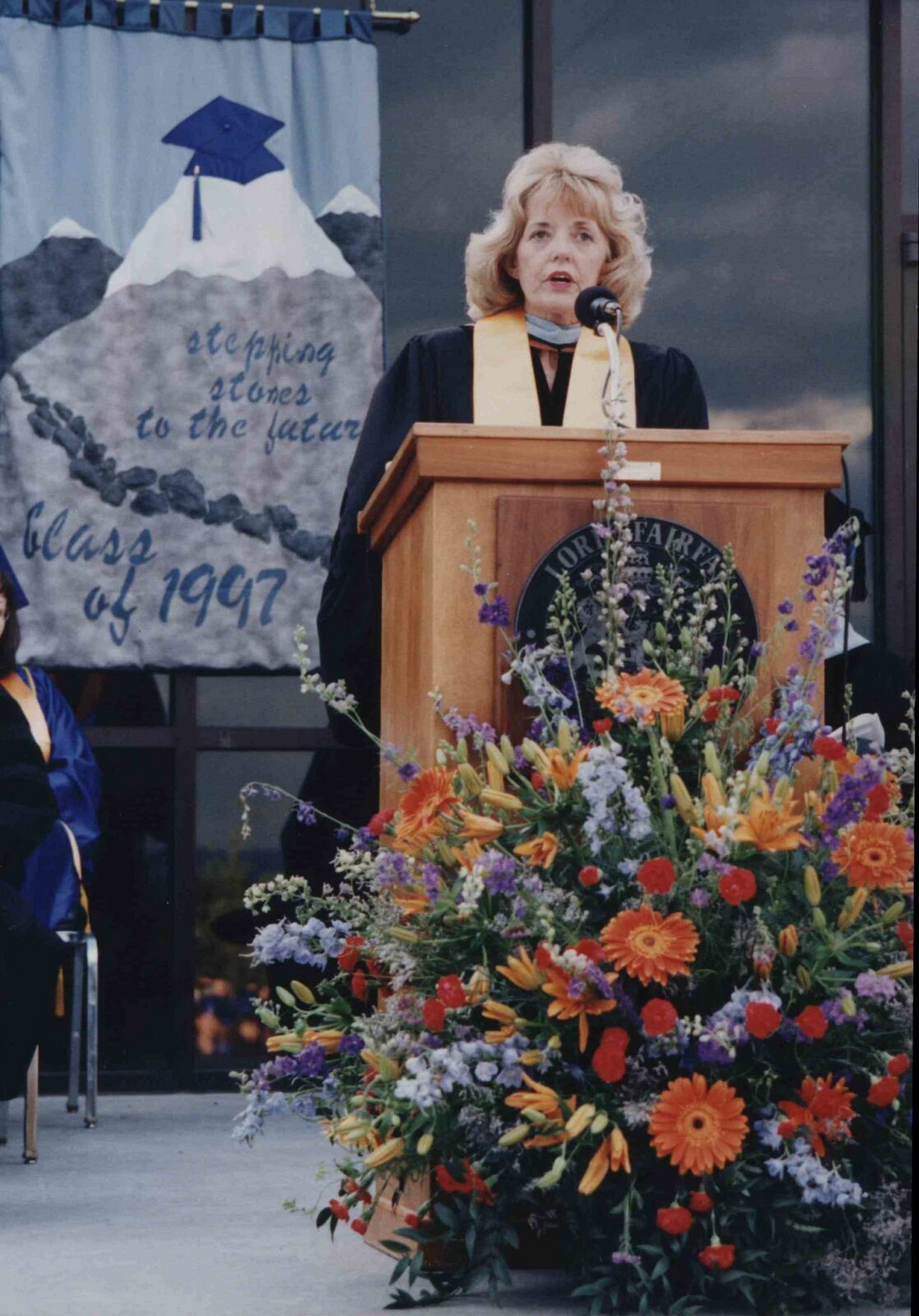 A woman with shoulder-length blonde hair speaks at a wooden podium, wearing a black academic gown with a gold stole. A large floral arrangement with orange, red, and purple flowers sits in front of the podium. To the left, a banner reads "Stepping Stones to the Future, Class of 1997" above an illustration of a mountain peak topped with a graduation cap. Another graduate in a blue gown is partially visible seated to the left.