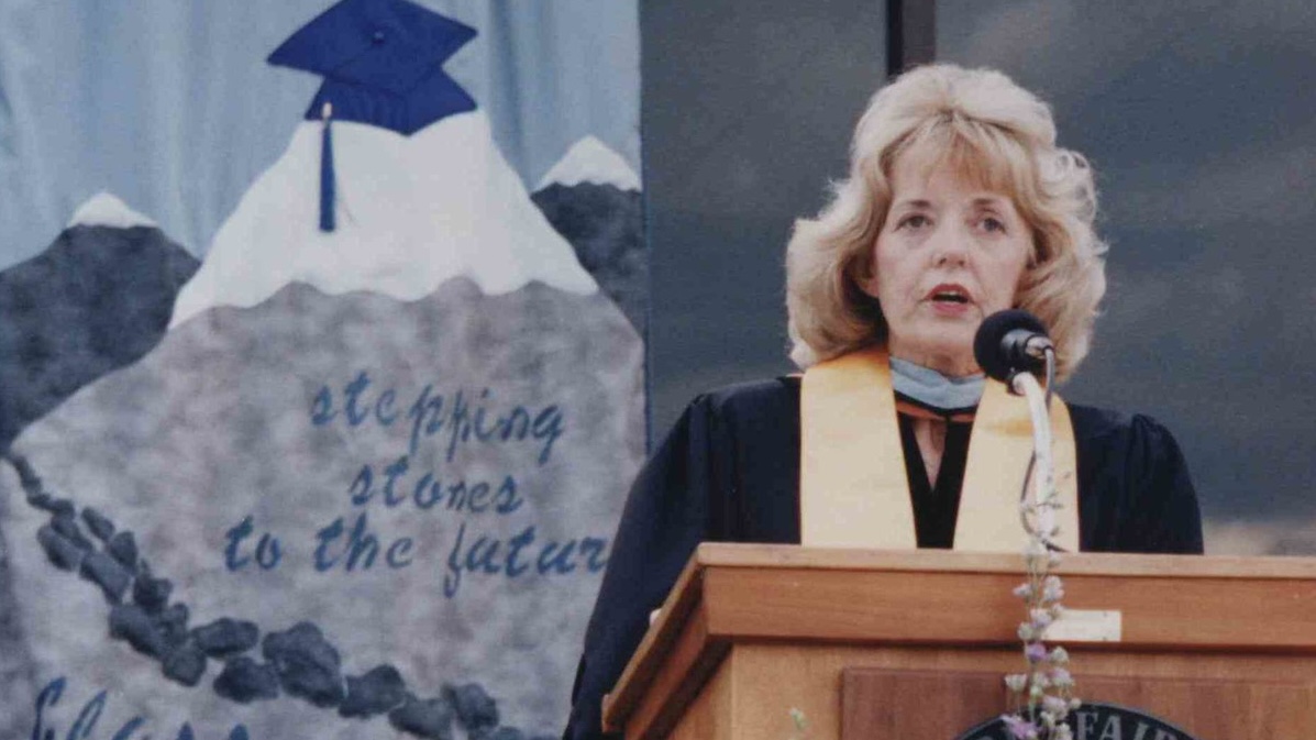 A woman with shoulder-length blonde hair speaks at a wooden podium, wearing a black academic gown with a gold stole. A large floral arrangement with orange, red, and purple flowers sits in front of the podium. To the left, a banner reads "Stepping Stones to the Future, Class of 1997" above an illustration of a mountain peak topped with a graduation cap. Another graduate in a blue gown is partially visible seated to the left.