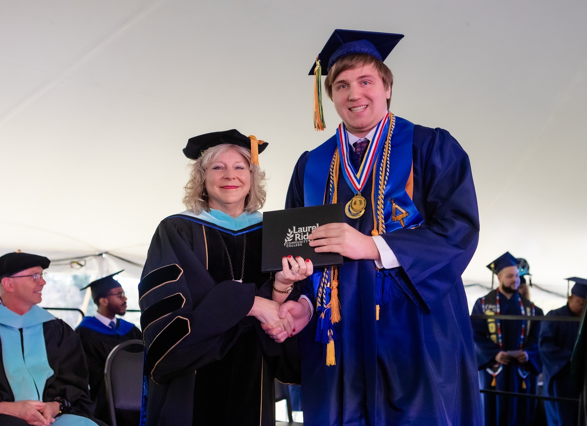 A Laurel Ridge Community College administrator in black doctoral regalia with light blue hood and gold trim shakes hands with a graduate in a blue cap and gown while together they hold a Laurel Ridge Community College diploma portfolio. The graduate wears multiple honor cords in gold and blue, a red, white, and blue medallion necklace, and an academic medallion, during an outdoor commencement ceremony held under a white tent.