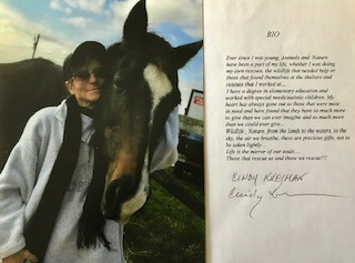 Artist Cindy Kreiman stands beside a horse outdoors, smiling as she embraces the animal.