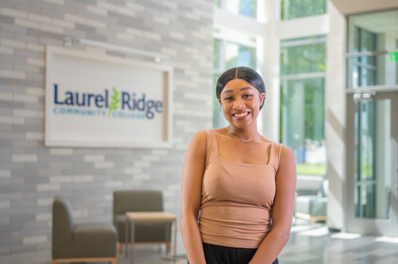A smiling female student with dark hair pulled back stands in a bright campus lobby, wearing a tan sleeveless top and a delicate necklace. Behind her, the Laurel Ridge Community College logo is mounted on a textured stone wall. Large windows and seating areas are visible in the background.
