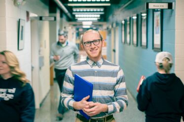 A smiling male student with short red hair and black-rimmed glasses holds a blue folder or laptop while standing in a campus hallway. He wears a striped button-down shirt. Other students and a person in a Laurel Ridge sweatshirt are visible in the background. A room sign reading 