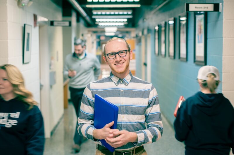 A smiling male student with short red hair and black-rimmed glasses holds a blue folder or laptop while standing in a campus hallway. He wears a striped button-down shirt. Other students and a person in a Laurel Ridge sweatshirt are visible in the background. A room sign reading "Room 216" is mounted on the teal-painted cinder block wall to the right.