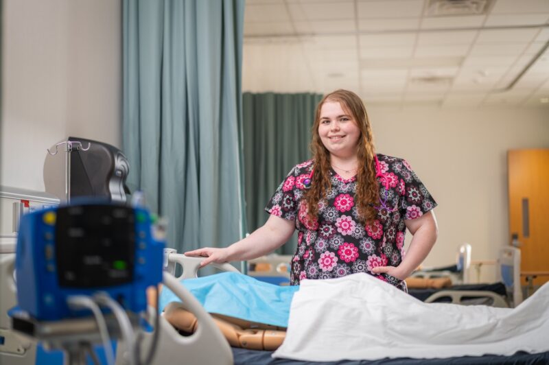 A smiling female nursing student with long auburn hair stands beside a hospital bed in a clinical simulation lab. She wears floral-patterned scrubs and has a stethoscope draped around her neck. A patient monitoring device and a mannequin partially covered by a white sheet are visible in the foreground.