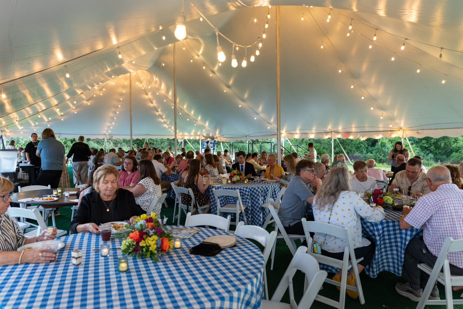 A large crowd of guests dine together under a white event tent decorated with strands of Edison bulb string lights. Round tables are covered with blue and white checkered tablecloths and adorned with colorful floral centerpieces featuring orange, yellow, and purple flowers alongside small votive candles. Guests seated in white folding chairs enjoy meals and conversation. Green trees are visible through the open sides of the tent in the background.