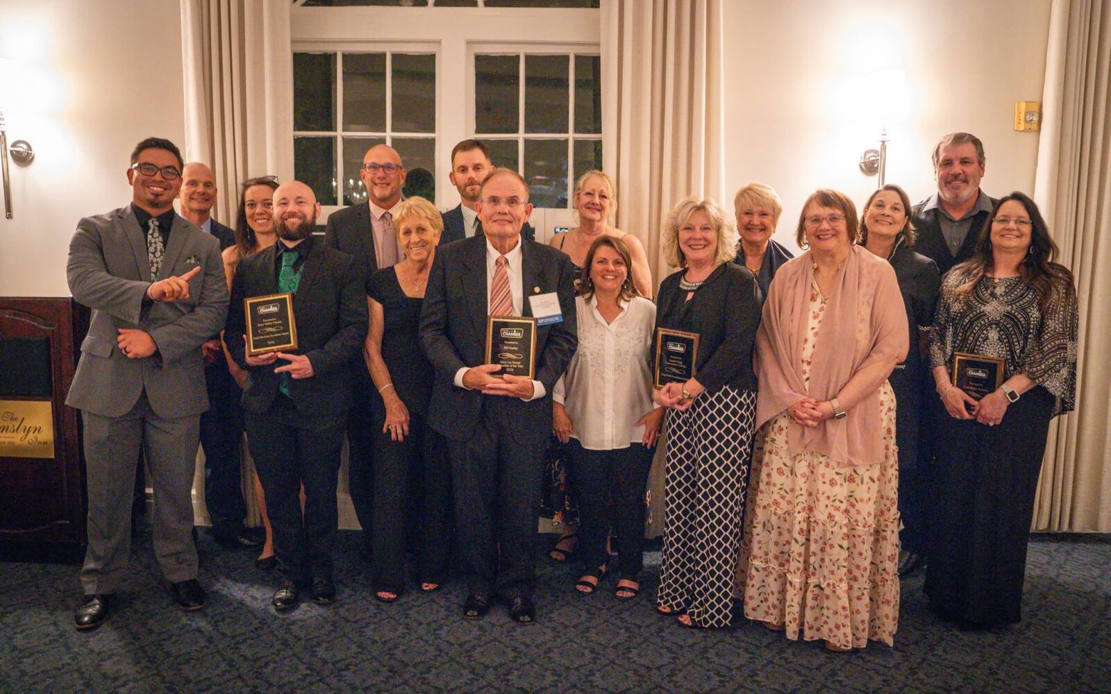 A large group of formally and semi-formally dressed adults pose together inside a banquet hall, smiling for a group photo. Several individuals in the front hold gold and black Chamber of Commerce award plaques. The setting appears to be an evening awards ceremony at The Mimslyn Inn, with warm ambient lighting, white-curtained windows, and carpeted flooring visible in the background.