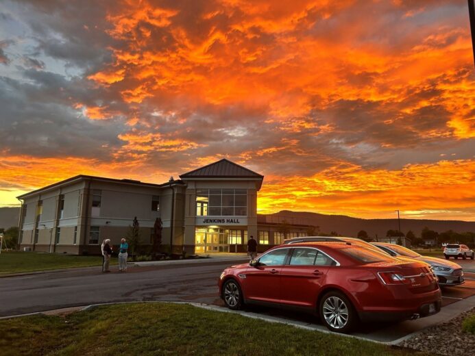 A dramatic sunset with vivid orange and red clouds fills the sky above Jenkins Hall at Laurel Ridge Community College's Luray-Page County Center. A red sedan and silver SUV are parked in the foreground, and two people converse near the building entrance. The illuminated lobby glows warmly against the darkening sky, with a mountain ridgeline visible in the background.