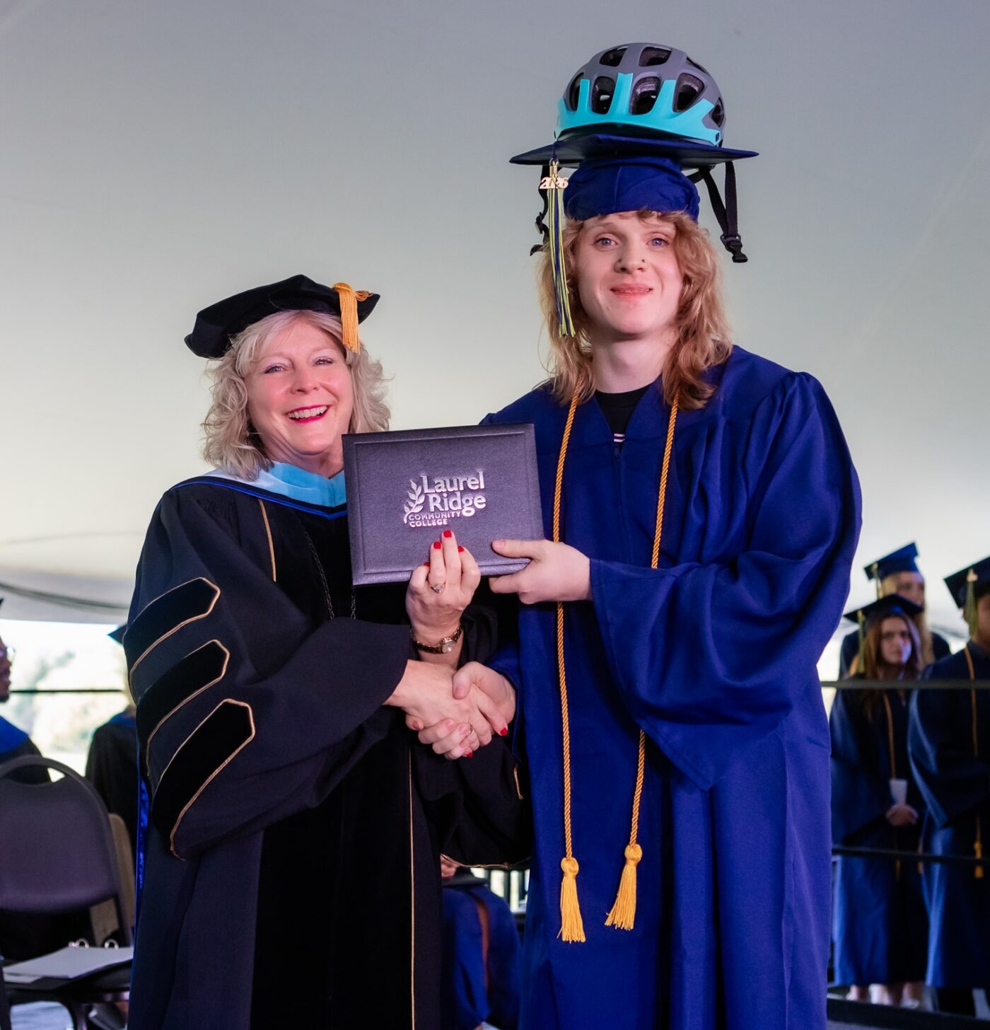 A Laurel Ridge Community College graduate wearing a bicycle helmet over his cap and blue graduation gown with gold honor cords shakes hands with a college administrator in academic regalia while receiving his diploma case under a white event tent, with other graduates seated in the background.