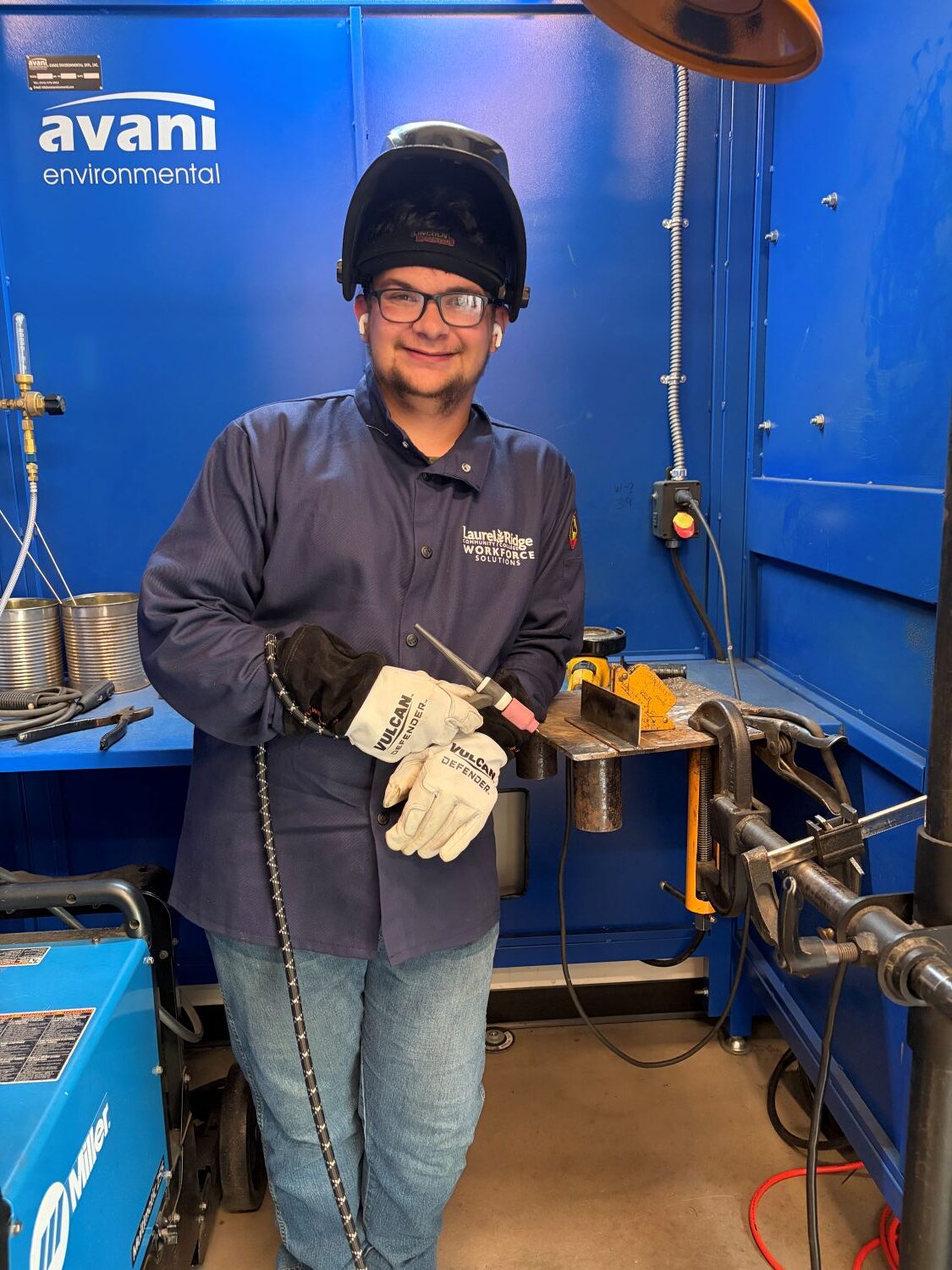 Matthew Del Vacchio stands smiling in a welding booth, wearing a black Lincoln welding helmet flipped up on his head, clear safety glasses, and wireless earbuds. He wears a navy Laurel Ridge Community College Workforce Solutions welding jacket over blue jeans and holds a TIG torch with white Vulcan Defender leather gloves. Behind him is a blue equipment panel with an "avani environmental" logo, a metal workbench with clamps and tools, and a blue Miller welding machine to his left.