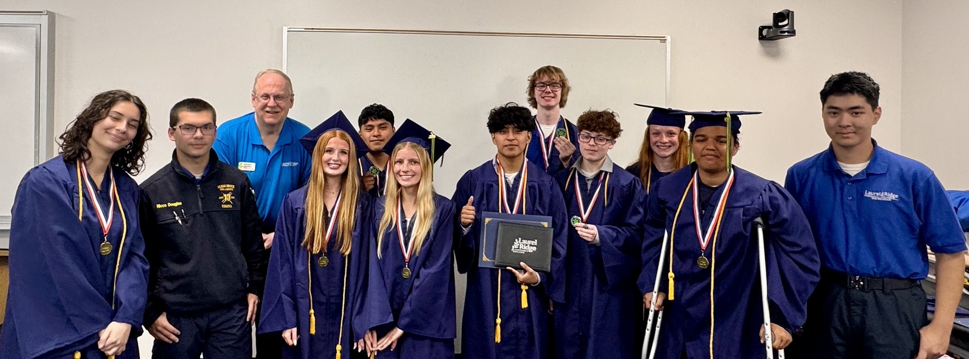 A group of graduates in navy blue caps and gowns pose together in a classroom, most wearing medals on red, white, and blue ribbons and honor cords in gold. In the center, a graduate holds a navy Laurel Ridge diploma folder while giving a thumbs up. A man in a blue Laurel Ridge polo shirt stands among the students toward the back, and a young man in a Laurel Ridge polo shirt stands at the far right. One graduate on the right stands with crutches. A whiteboard and mounted camera are visible on the wall behind the group.
