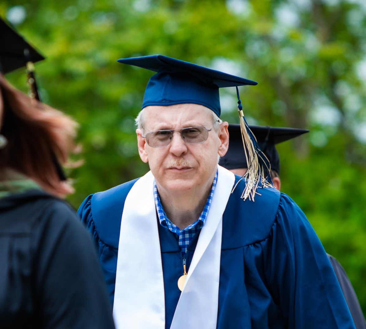 A man in a navy blue cap and gown walks outdoors at a graduation ceremony, wearing glasses, a blue and white checkered collared shirt, a white stole, and a gold medallion on a cord around his neck. His cap has a gold tassel. Blurred greenery fills the background, and other graduates in caps and gowns are visible around him.