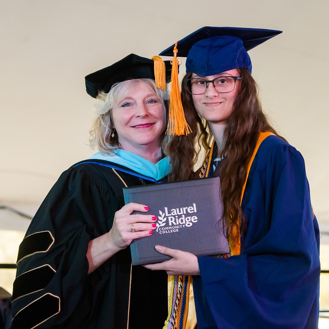Rachel poses for a photo at a Laurel Ridge Community College commencement ceremony. She wears a navy blue graduation cap and gown with a gold tassel, honor cords, and a stole, and has long brown hair and glasses. Standing beside her is a faculty member or administrator with blond hair, dressed in black doctoral regalia with velvet trim and a teal hood. Together they hold a gray diploma cover displaying the white Laurel Ridge Community College logo with a leaf design. They stand under a tent against a cream-colored backdrop.