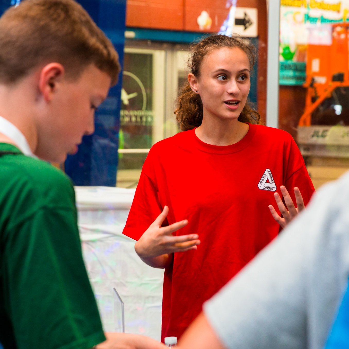 Rachel Stange, a young woman with curly brown hair pulled back into a ponytail, wears a red t-shirt with a triangular chest logo reading "INFOTECH CYBERSEC COMPSCI." She gestures with both hands while speaking to a group at an indoor event. In the foreground, a boy with short reddish-brown hair in a green shirt looks down at a table, and another person in a light blue shirt stands partially visible on the right. Behind her, glass storefront windows, colorful signage, and a black directional arrow are visible.