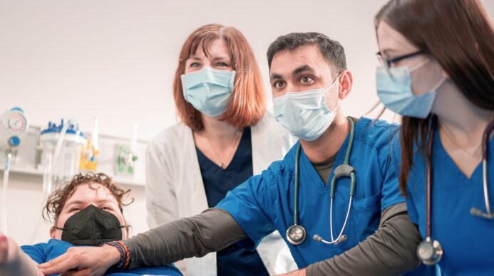 Four nursing students and an instructor gather closely around a patient bed in a clinical simulation lab, all wearing surgical face masks. An instructor in a white lab coat over navy scrubs stands at center. Beside her, a student in blue scrubs with a stethoscope around his neck reaches forward toward the simulation manikin, while another student in blue scrubs and glasses leans in from the right. A fourth student in blue scrubs and a black mask crouches at the lower left. Medical equipment including a suction canister and wall-mounted gauge is visible in the background.