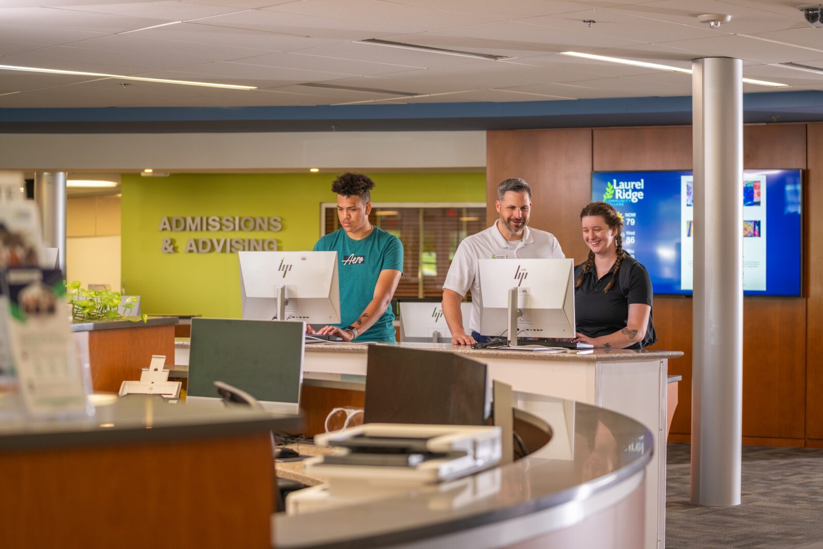 Three people stand at a service counter in the Laurel Ridge Community College Admissions and Advising office. A young man in a teal Aero T-shirt works at an HP computer on the left, a smiling staff member in a white polo shirt stands in the center with his hands resting on the counter, and a young woman with a braided ponytail in a black polo shirt smiles as she works at another HP computer on the right. Behind them, large silver letters on a lime-green wall read "Admissions & Advising," and a blue digital display shows the Laurel Ridge Community College logo alongside the local weather forecast. The modern lobby features wood-paneled accents, a potted plant, and a rounded reception desk in the foreground.