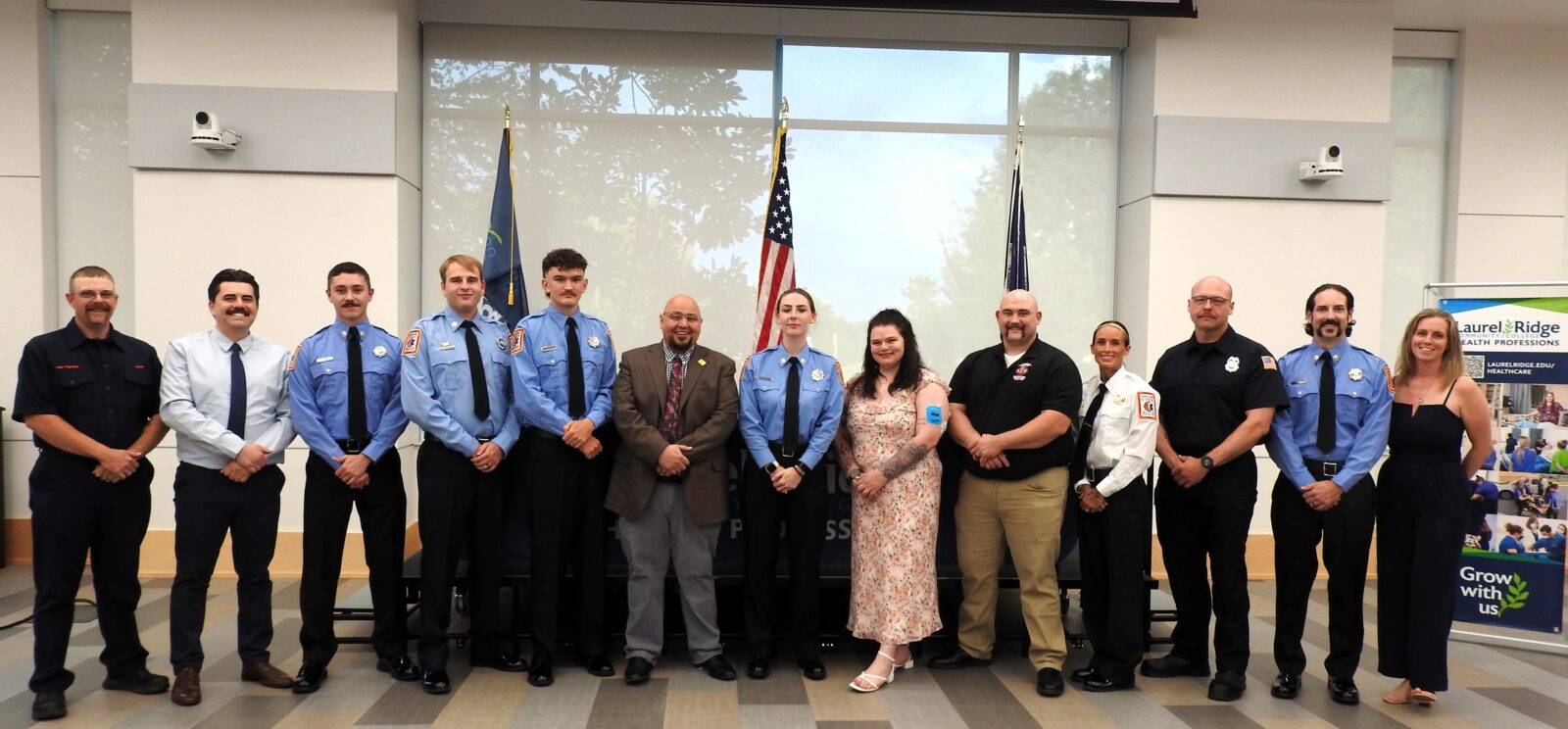 Group photo of paramedic program graduates, EMS staff, and Laurel Ridge Community College representatives posing together at the Hybrid Paramedic Program Graduation Ceremony, with American and organizational flags in the background.